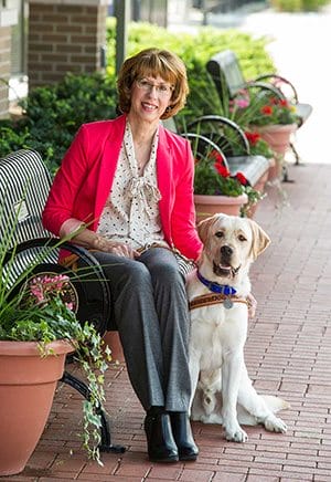 Sue Daniels sits outdoors on a bench with a yellow lab seated next to her on the ground. Sue has one hand on the dog