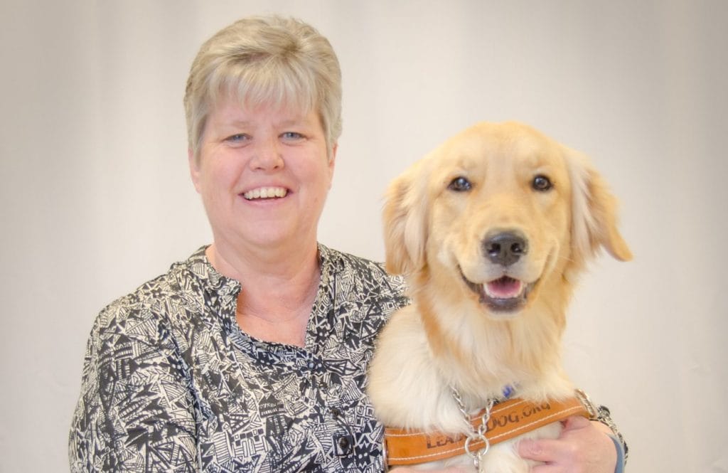 A woman wearing a flowery button up shirt has her arm around the golden retriever in Leader Dog harness.
