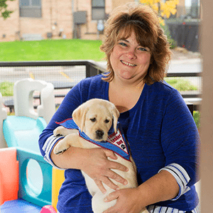 Photo of Bev smiling at the camera and holding a yellow lab puppy, who is wearing a blue Future Leader Dog bandanna