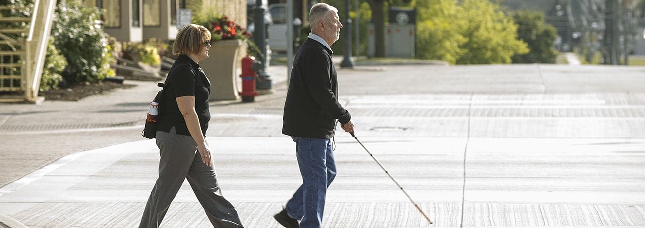 O&M Client2 A man crosses and intersection. He is traveling with a white cane in his right hand and is followed by a female certified orientation & mobility specialist