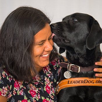 Close-up photo of a woman wearing a flowery shirt next to a black lab in leather Leader Dog harness. The woman is smiling and her arm is around the dog. The dog is licking her cheek