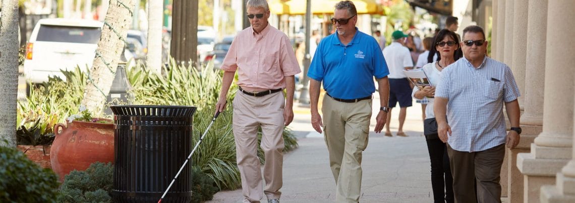 A man with a white cane walks down a busy sidewalk with another man wearing a Leader Dog polo