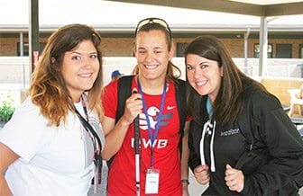 Three young women stand outdoors facing the camera, smiling. The woman in the center is holding a white cane