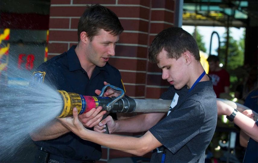 A fireman assists teenager Brock with holding a fire hose that is spraying water