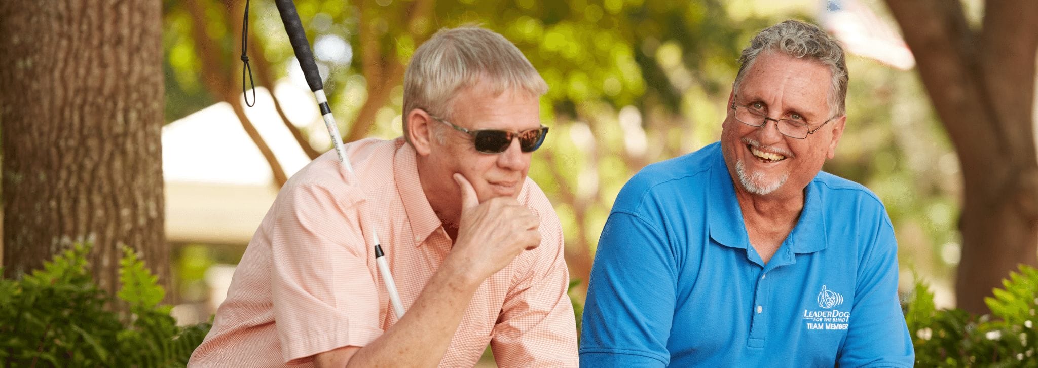 Two men sit side by side. One, wearing an orange shirt and sunglasses leans forward with his hand resting on this chin and his white cane leaning against his shoulder. The other smiles, wearing a turquoise Leader Dogs for the Blind polo.