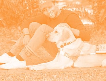 Orange colored image of a young man sitting on a sidewalk. He is smiling and petting the dog next to him in harness. The dog is lying down next to the man