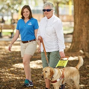 Sarah Duyck Two women, one older and one younger, walk outdoors on a sunny day. The older woman is walking with a yellow lab guide dog in harness. The younger woman is beside them, smiling and observing