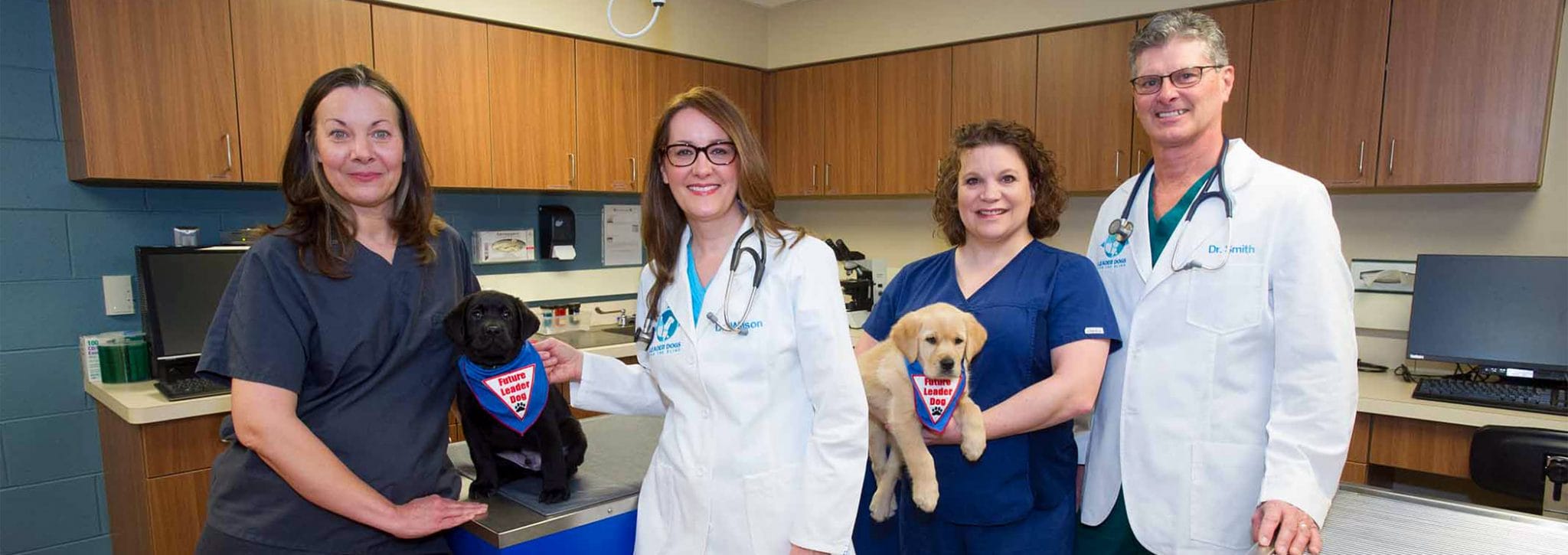 Three women and one man stand facing and camera and smiling in our brightly lit vet clinic. With them are two lab puppies, one black and one yellow, wearing Future Leader Dog bandannas