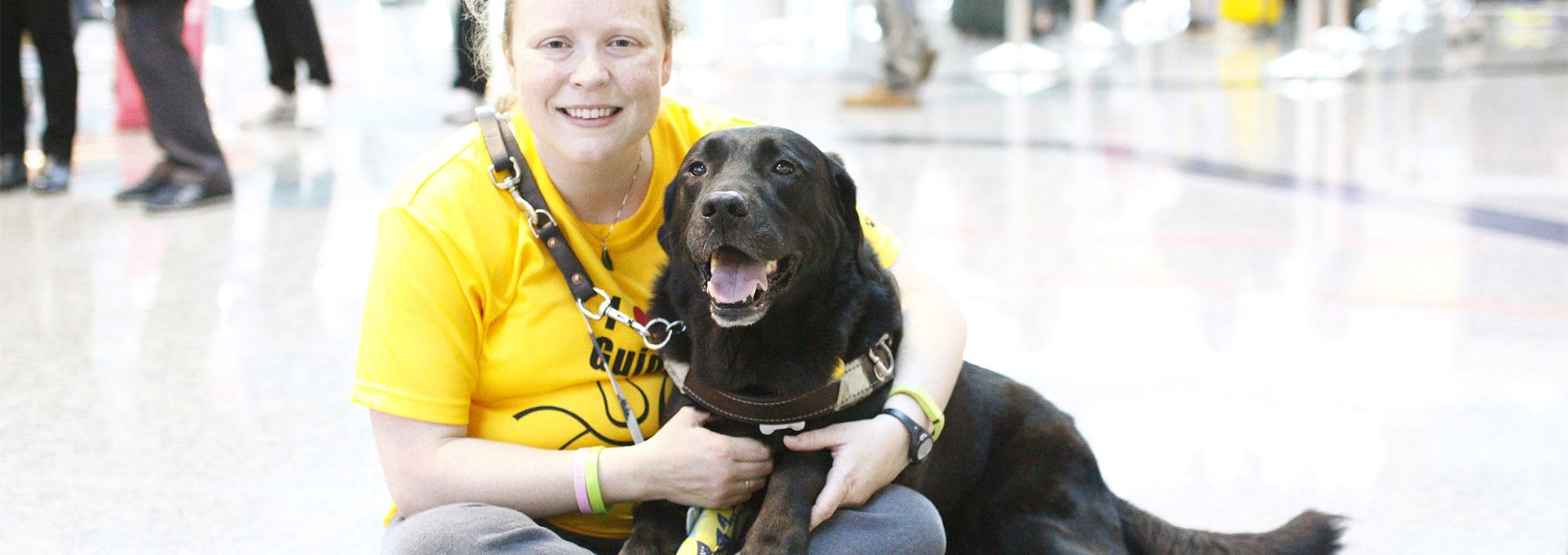 A woman sits cross-legged on a white floor with a black lab in harness partially on her lap. She is smiling at the camera