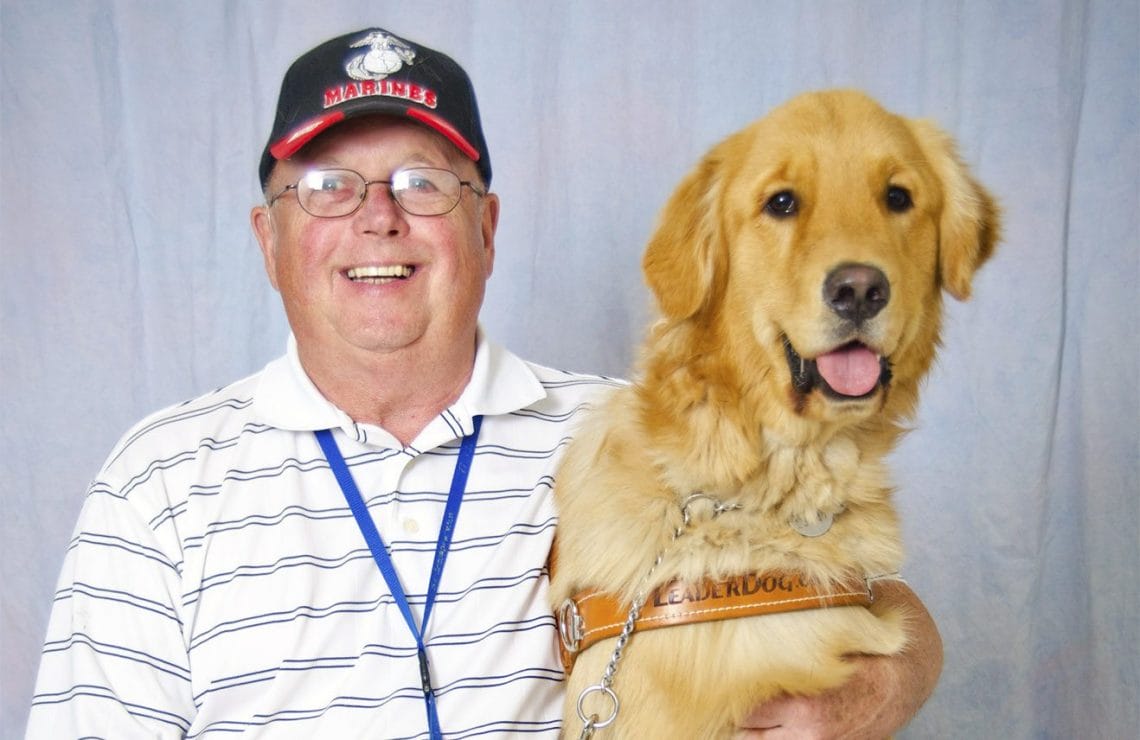 Bob Justin sits in front of a gray background wearing a black and white striped polo shirt and a Marines cap. Next to him in Leader Dog harness is golden retriever Lego