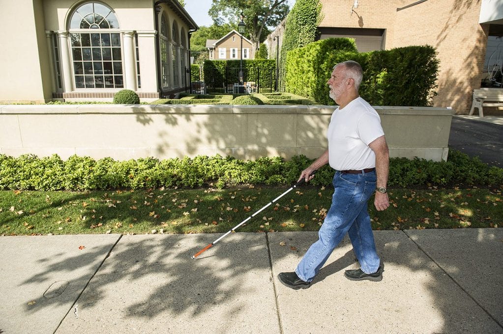 A man walks down a sunny, grass-lined sidewalk using a white cane