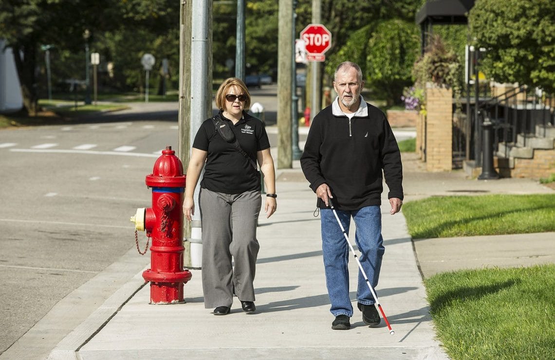 Bob Rock uses a white cane to navigate down a sidewalk on a sunny day. Slightly behind him is a woman in a Leader Dog polo observing his progress