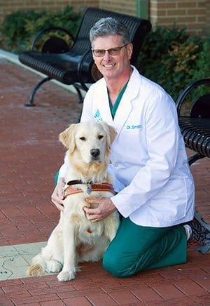 Dr. Smith kneels on brick while wearing scrubs and his white coat. He is smiling at the camera with his hands on a golden retriever in harness sitting next to him
