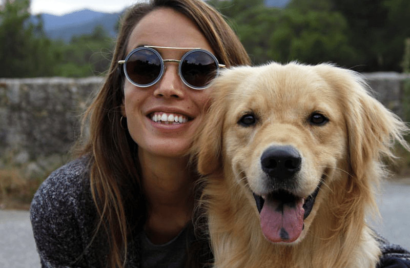 Close up of a young woman wearing sunglasses and smiling at the camera. In front of her and just to her left sits a golden retriever with his mouth open as if smiling