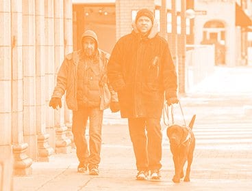 Two men, one with a guide dog in harness, walk down a sidewalk in winter