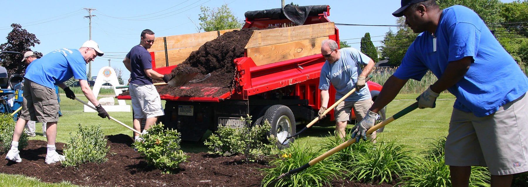 Four men, each with a rake or shovel, stand outdoors by a truck dumping soil onto a garden bed. The men are spreading the soil around