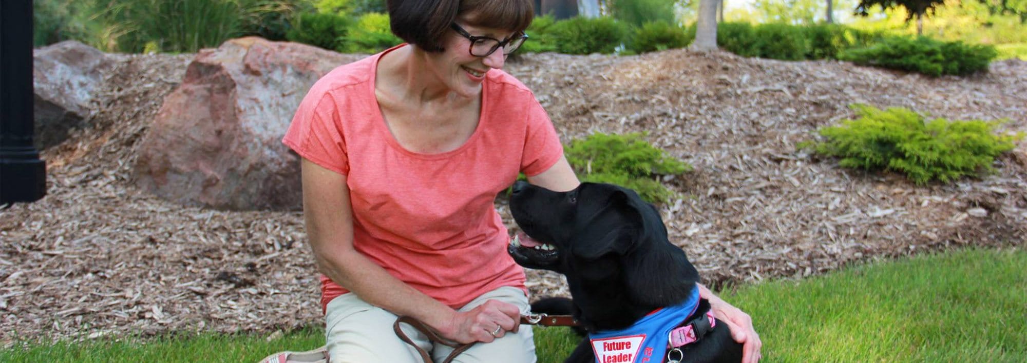 A woman kneels on grass in a neatly manicured yard. She is smiling at the black lab lying next to her on the grass. The lab is wearing a Future Leader Dog bandanna and looking up at the woman