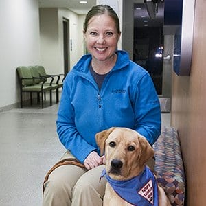 Laura sits on an indoor bench in a hallway smiling at the camera and holding the lash of the young yellow lab in Future Leader Dog bandanna. The lab is sitting in front of Laura's legs