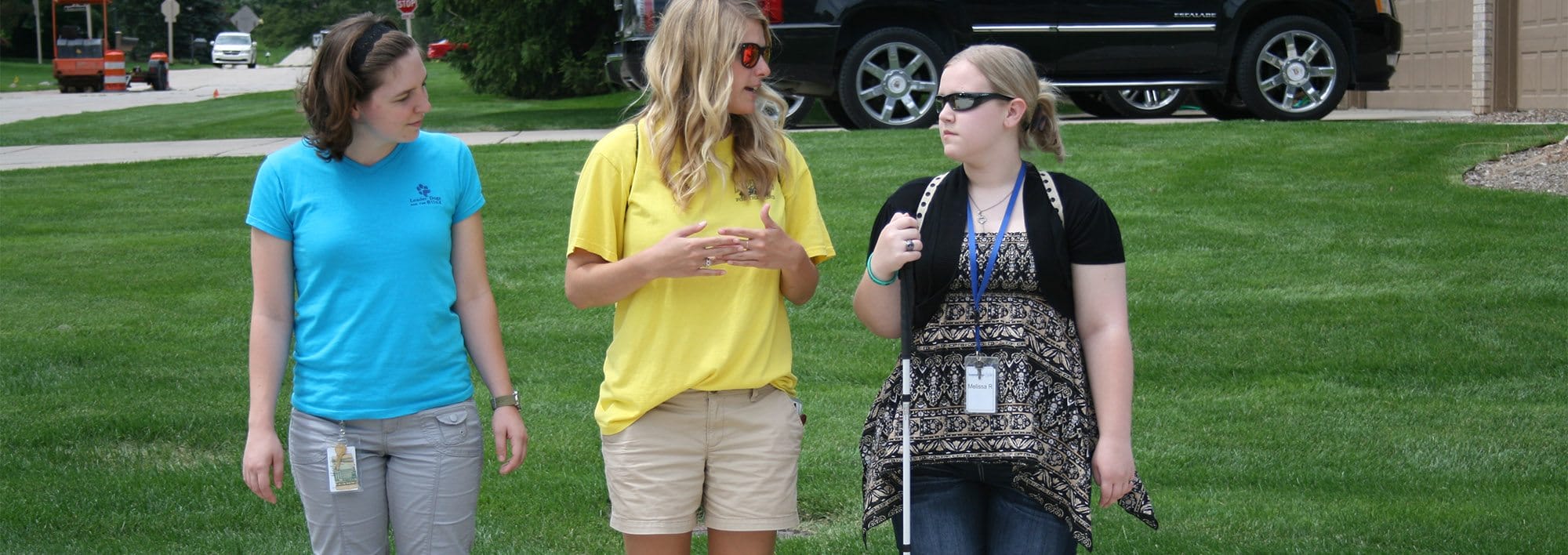 Three young woman stand outdoors in front a lawn. One woman is holding a white cane while the middle woman appears to be talking