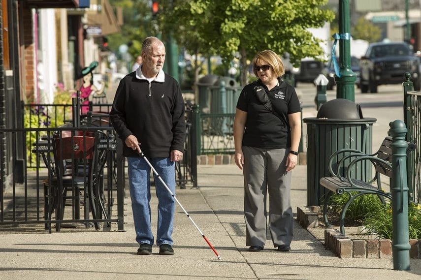 A man walks down a sunny sidewalk with outdoor tables. He is walking with a white cane next to a woman in a Leader Dog polo