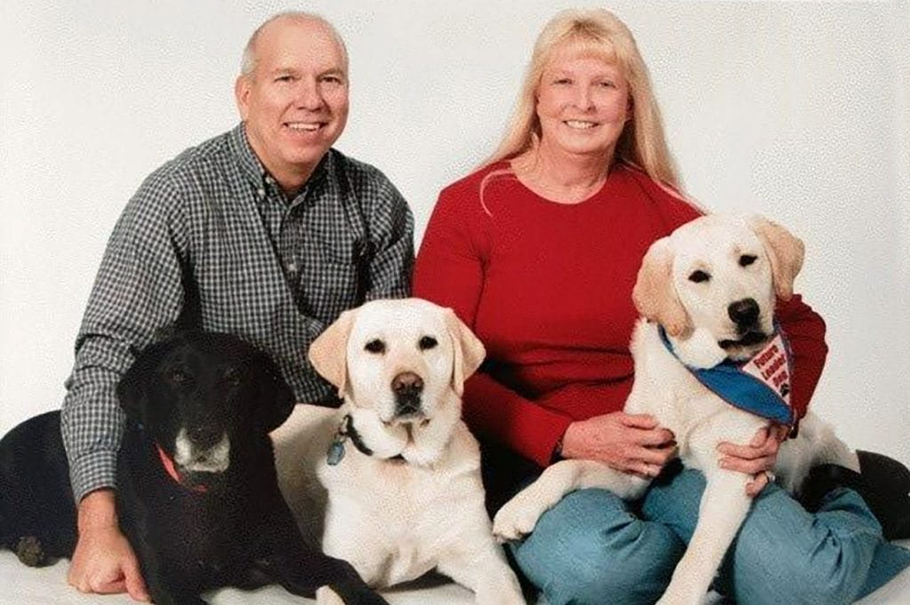 Glenn and Ginger Rossow sit on the floor in front of a white background with three Labradors, two yellow and one black, lying in front of them