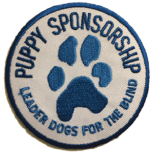 A white circular patch with a blue border. The patch has a blue paw print embroidered in the middle and surrounded by the words "Puppy Sponsorship" and "Leader Dogs for the Blind"