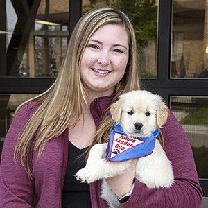 Sam stands in front of the glass garage doors in the Canine Center smiling at the camera and holding a young golden retreiver puppy with a Future Leader Dog bandanna on