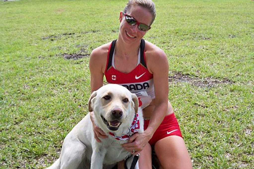 Noella sits outdoors in athletic gear, smiling at the camera with her arms around Leader Dog Zeke, who sits in front of her