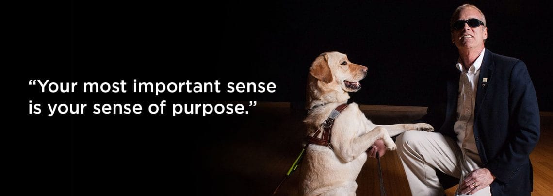 Platzer with quote A man wearing a suit and sunglasses kneel on a wood floor. He is smiling toward the camera and holding the paw of a yellow lab in harness that is sitting on its hind legs. The background is black and has a quotation in white reading "Your most important sense is your sense of purpose."