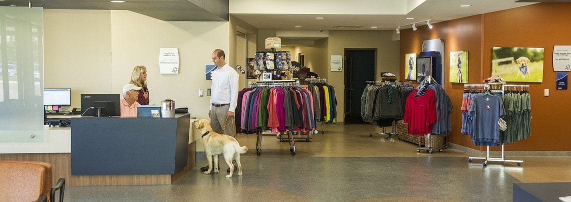 The bright, open space of the canine center lobby on Leader Dog's campus. Racks of clothing can be seen in the background. A man and a dog stand near the lobby desk, where another man and woman are conversing