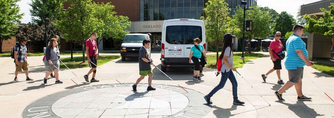 A group of people, six with white canes and two without, walking parallel to the camera across part of Oakland University's campus on a sunny summer day