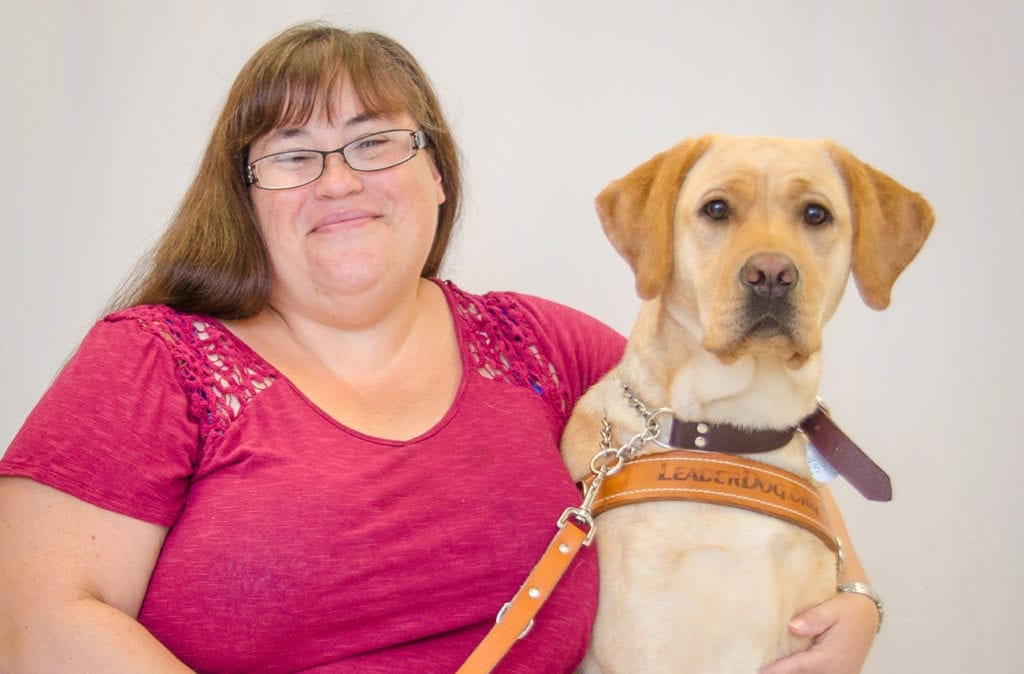 Amy Chevalier sits facing the camera and smiling with her left arm around yellow Labrador Leader Dog Ember, who also sits facing the camera in harness