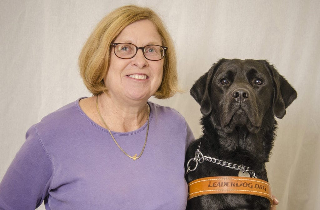 Gretchen Preston sits smiling at the camera with her left arm around her Leader Dog, black Labrador Floyd, who is wearing a leather Leader Dog harness