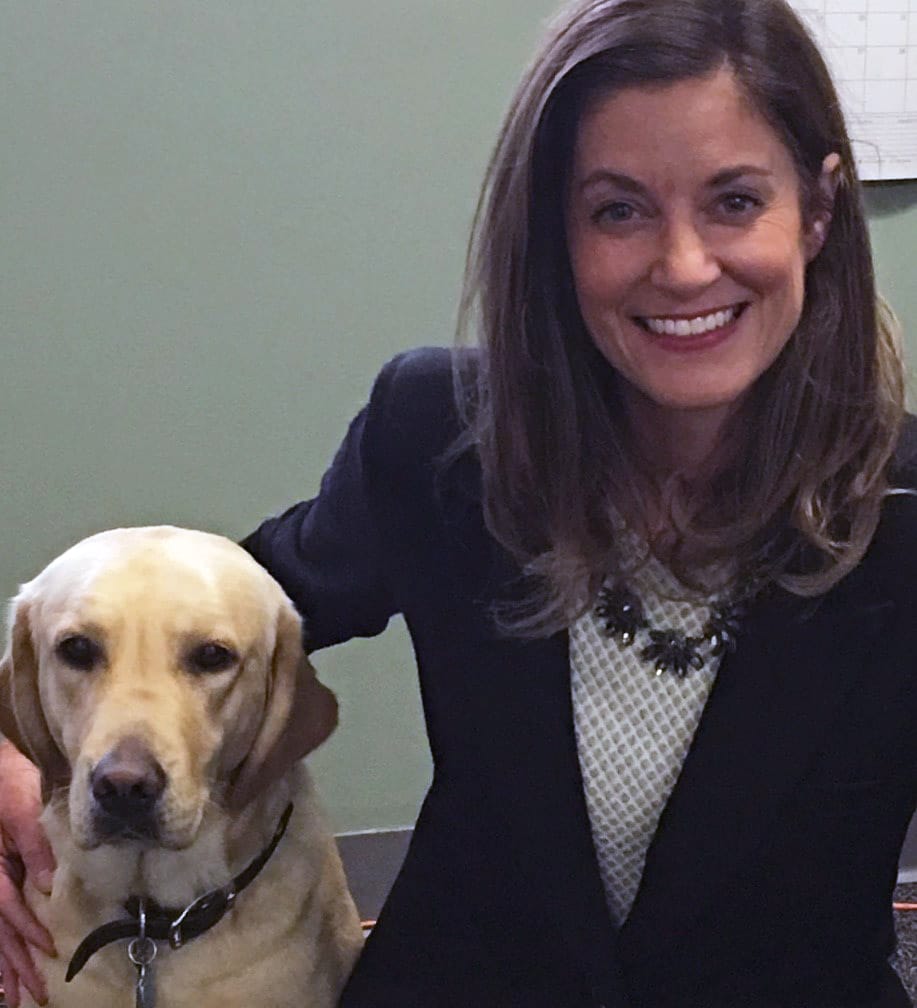Melissa crouches, smiling, with her arm around a yellow Labrador sitting next to her