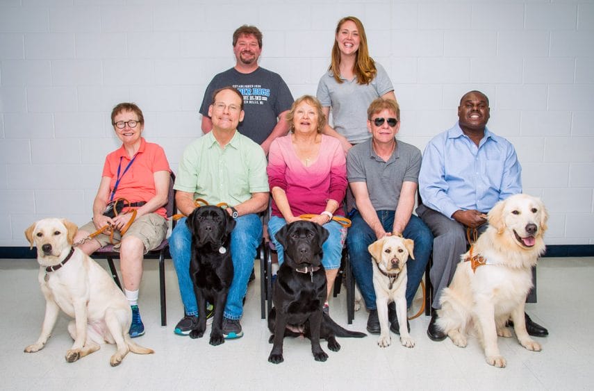 Five clients are seated, facing the camera and smiling. Each one has a Leader Dog in harness sitting on the floor next to them. Behind the clients stand two instructors, one male and one female