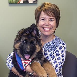 Beth sits on a chair indoors, smiling at the camera and holding a fluffy German shepherd puppy that is wearing a blue Future Leader Dog bandana