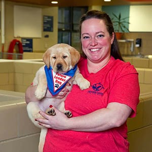 Dana stands in the canine center smiling and holding a yellow Labrador puppy that is wearing a blue Future Leader Dog bandanna