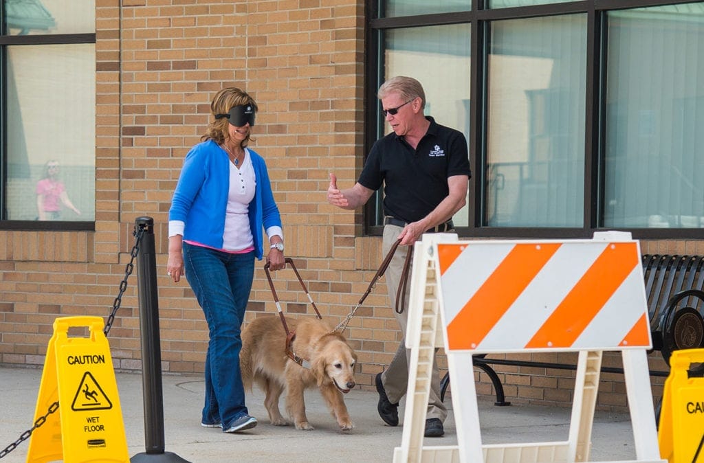 A woman wearing a blindfold walks toward some roadblocks while holding the handle of a harness on a golden retriever. The golden retriever is guiding the woman while a Leader Dog instructor holds the dog's leash and speaks to the woman