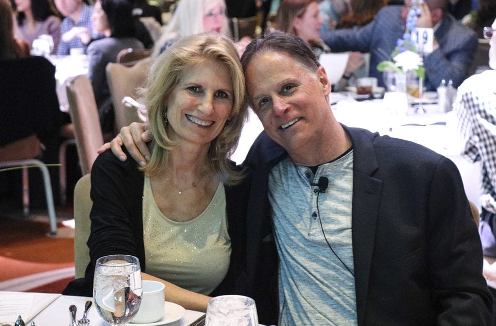 Jeff and his wife, Linda, sit with their arms around each other's shoulders, smiling at the camera. They are seated at a table with other tables and guests visible in the background.
