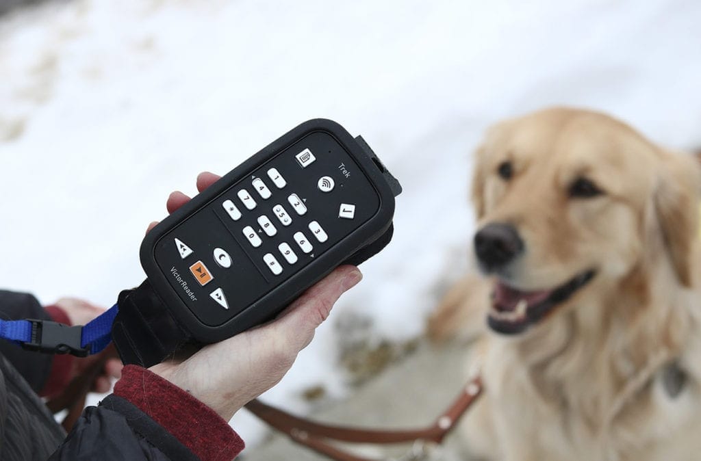 Close-up photo of a person holding a Victor Reader Trek GPS device. In the background is a golden retriever looking up toward the person