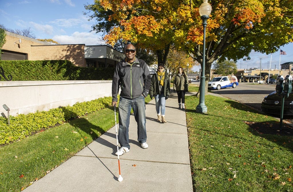A man walks down a sidewalk on a sunny day. He is using a white cane and smiling