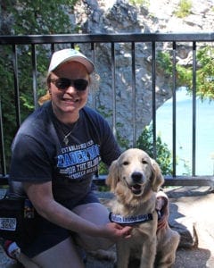 An adult female puppy raiser kneels next to a golden retriever puppy in its blue Future Leader Dog vest. They are outdoors in front of a rocky outcropping