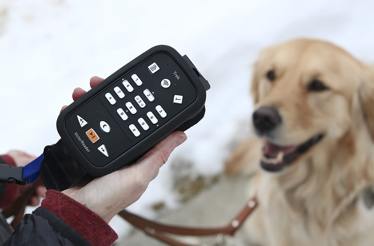 Close-up of a hand holding the Victor Reader Trek GPS device. In the background is snow and a golden retriever is visible, looking up at the person with the GPS