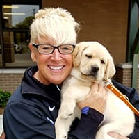 Laura Banks holds a young yellow lab puppy in her arms, smiling at the camera