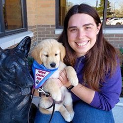 Kayla crouches next to a metal German shepherd statue, smiling and holding a golden retriever puppy wearing a Future Leader Dog bandana