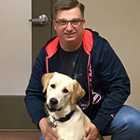 Mike Pikelis kneels next to a sitting yellow lab