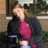 Krista Stauffer kneels next to a black lab that is licking her nose