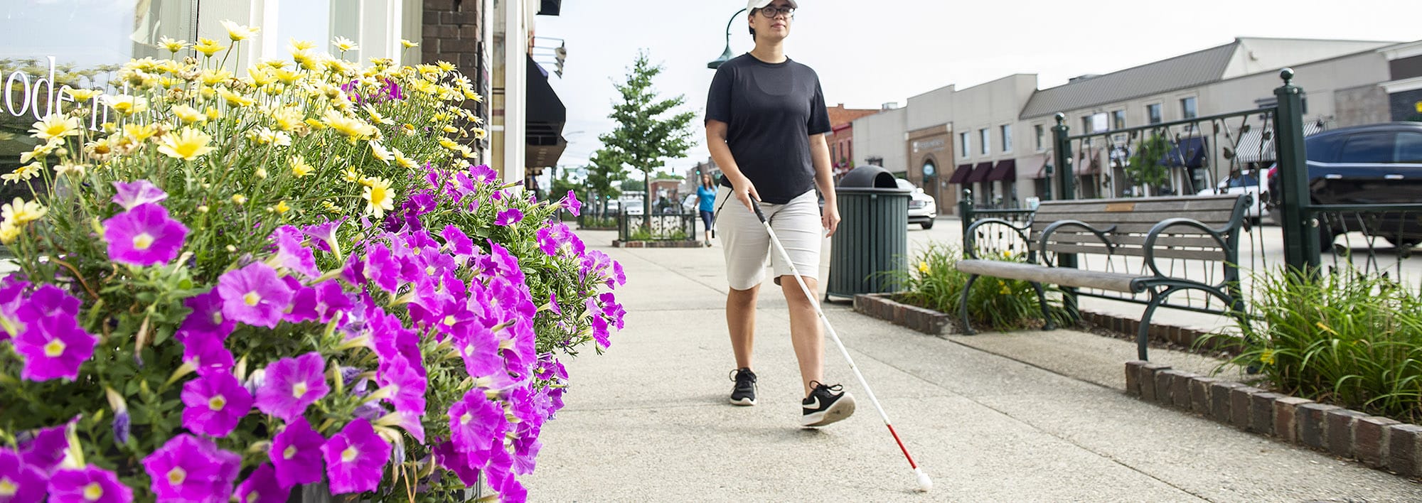 O&M Client Brooke Coleman A young woman walks along a downtown sidewalk with traffic on her left side and flower boxes and a storefront on her right. She is using a white cane to navigate