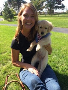 Ashley sitting on grass on a sunny day at Leader Dog's campus. She is smiling and holding a golden retriever puppy wearing its blue Future Leader Dog bandana.