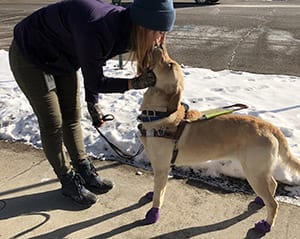 Ashley stands outside on a sidewalk. There is snow next to the sidewalk. She is leaning over a golden retriever in harness and giving it a kiss on the nose.